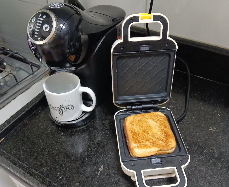 Foto de um sanduíche com uma casca dourada dentro de uma sanduicheira elétrica. Ao lado tem uma máquina de café e uma xícara escrito Canastra